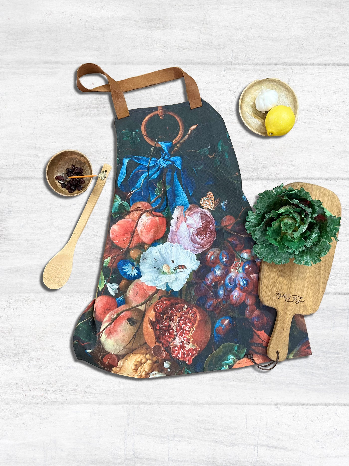 Apron with leather straps laid out on light wood table. Surrounded by cookwares, utensils and vegetables like garlic, lettuce, lemon and raisons. The Apron is printed showing artwork titled Still Life. The print shows various fruits and flowers in vibrant red, orange and blue hues against a dark background. There are insects in open fruit and crawling over flowers.