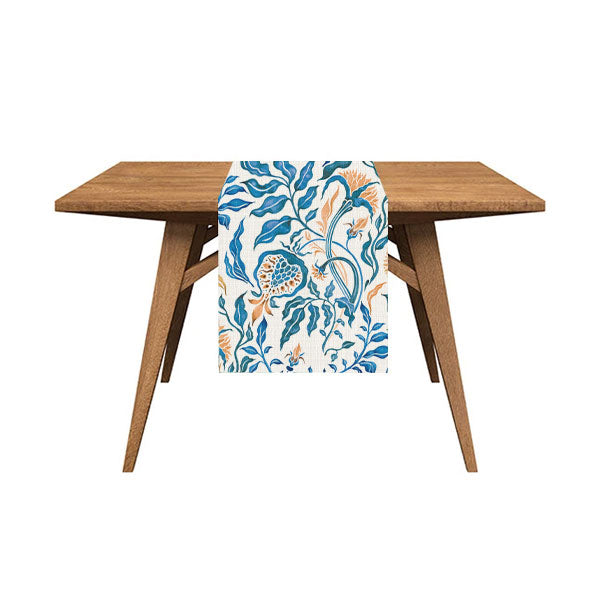 square table side view on white background. Mockup image showing runner on table and hanging halfway down. Runner has a blue and mustard yellow patterns of illustrated leaves and flowers. The leaves and stalks of the flowers are blue while the buds are yellow